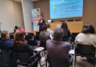 Dans une salle, face à un écran et un conférencier, des personnes assistent à une présentation