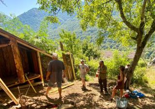 Quatre personnes se tiennent aux abords d'une construction de bois, dans un paysage de collines boisées