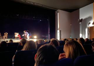 Dans une salle de spectacle, sur scène, une vieille femme très courbée