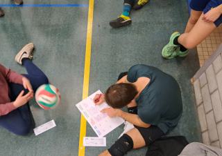 Une personne assise sur le sol rempli un formulaire, vu d'en haut, avec un ballon de volley ball lancé vers la personne 