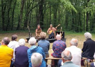 Un public assis sur des bancs dans une forêt, regardent deux musiciens qui leur font face