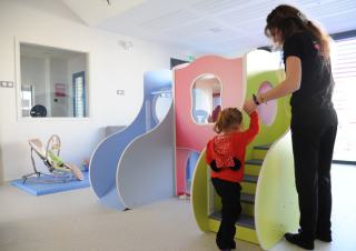 Une jeune femme aide une petite fille à monter les escaliers d'une structure de jeux, dans une salle aux murs blancs