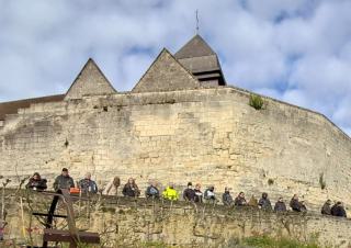 Au pied du mur d'enceinte d'un château, des personnes se tiennent contre un muret