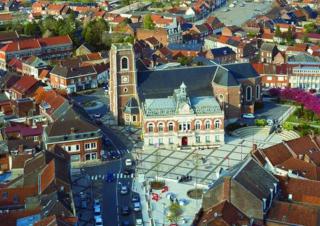 Vue du ciel d'un village de brique rouge. Au centre, un édifice religieux de brique rouge et un parvis a rayures obliques