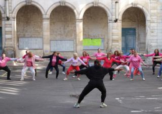 Des femmes vêtues de rose se tiennent en ligne devant une personne vêtue de noir et reproduisent ses mouvements. Au fond, des arcades.