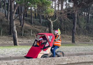 Deux personnes installées dans la cabine d'un vélo triporteur, et une personne qui pédale sur le vélo, sur une route en contrebas d'une forêt de pins