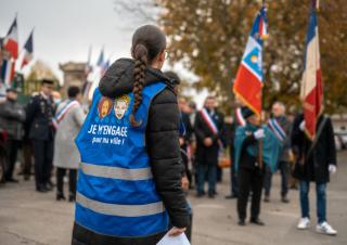 Cérémonie officielle avec drpeaux. Au premier plan une jeune femme de dos, longs cheveux tressés, gilet bleu sur lequel on peut lire "je m'engage pour ma ville"