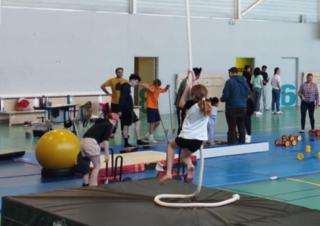 Dans un gymnase, au premier plan, on voit une jeune fille grimper à la corde