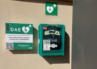 Sur un mur, un boitier de défibrilateur et un autre boitier, sur lequel on peut lire: une victime d'arrêt cardiaque a besoin de ce défibrilateur