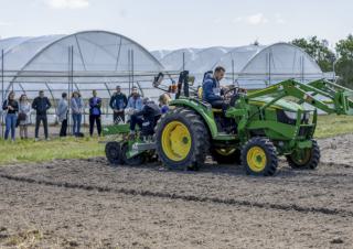 Un tracteur sur un champ labouré, en fond des personnes regardent le tracteur, elles sont devant des serres de culture