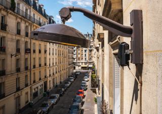 Une tête de lampadaire fixée à un bâtiment  dans rue parisienne. Sous la lampe, un boitier