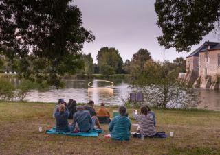 Des personnes assises devant une étendue d'eau sur laquelle flotte une forme sphérique. Au loin, un château.