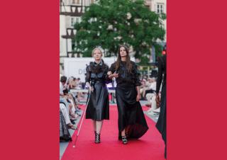 Deux femmes élgantes marchent sur un tapis rouge entouré de public. L'une d'entre elle se dirige avec une canne blanche