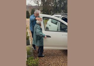 Un monsieur tient une portière de voiture pour laisser s'installer une personne aux cheveux blancs sur le fauteuil passager d'une voiture