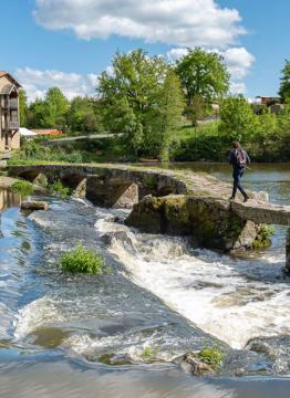 Une personne chemine sur une passerelle de pierre au dessus d'une rivière, en direction d'un moulin sur la berge opposée